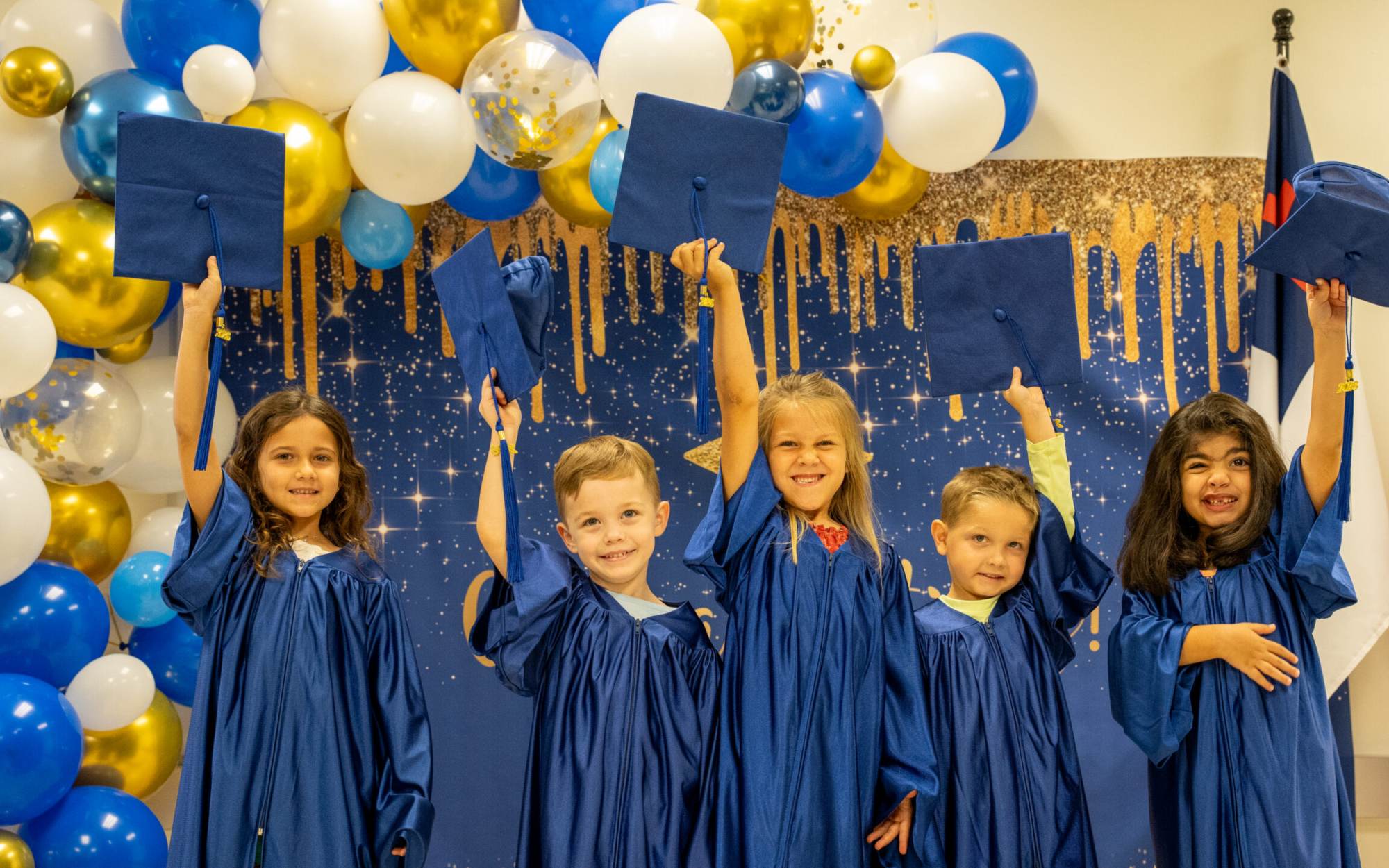 Preschool graduates in blue gowns holding their caps up high