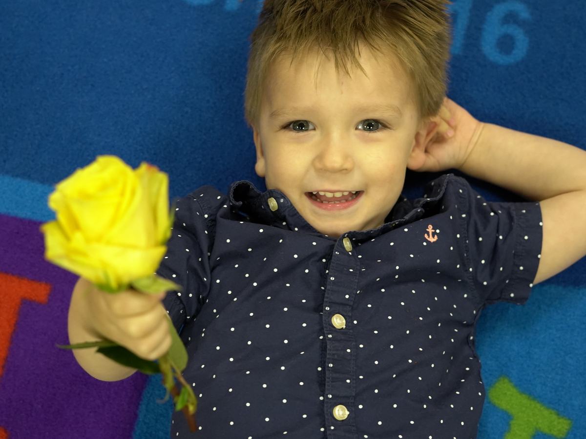 A grinning little boy lying on the classroom rug, holding out a yellow rose at Grandparents Day