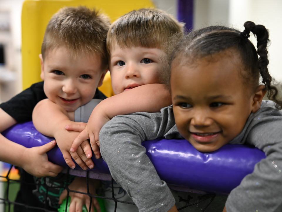 Three smiling children leaning over a padded rail in the indoor play room