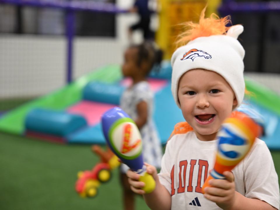 A laughing toddler in a fuzzy hat shaking two toy maracas