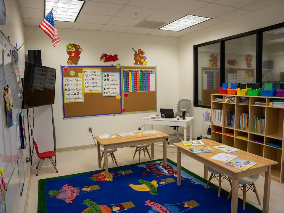 A classroom with wooden tables, a fruit-print rug, a bulletin board and bookshelves