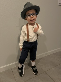 Santiago smiling in a grey fedora, blue glasses and brown suspenders, pointing to himself
